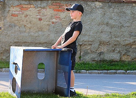 Ein junger Junge steht neben einem blauen und grauen Objekt, möglicherweise einem Verkaufsautomaten, am Grasrand der Straße.