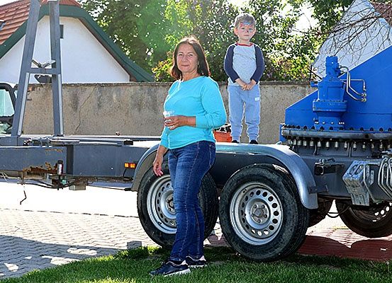 Eine Frau und ein Kind stehen neben einem blauen Anhänger. Die Frau hält ein Getränk in der Hand. Das Kind hat Farbe im Gesicht. Sie stehen vor einem Haus mit rotem Dach.