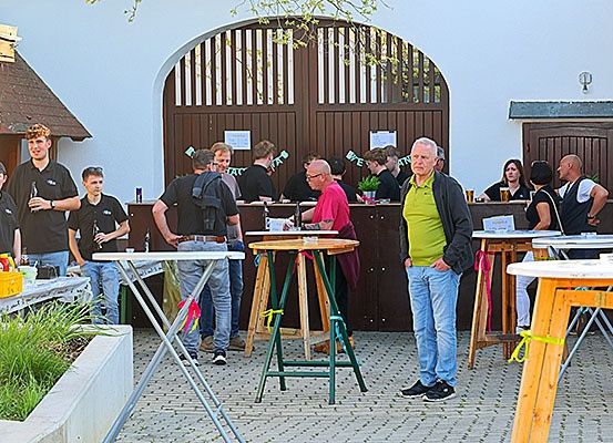 Menschen versammeln sich um Tische vor einem Gebäude. Einige trinken, andere stehen. Ein Mann in einem grünen Shirt hat die Hände in den Taschen.