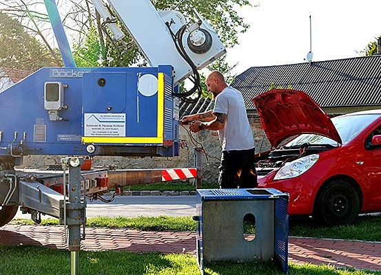 Ein Mann steht neben einem roten Auto mit offenem Motorhaube, möglicherweise arbeitet er daran. Ein blauer Kran ist am Straßenrand geparkt.