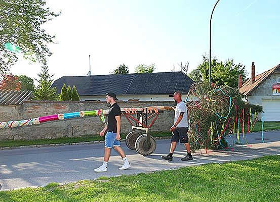 Zwei Männer gehen mit einem langen, bunten Objekt auf einer Straße und ziehen es hinter sich her. Dahinter steht ein Haus mit einem Schornstein und ein Auto, das davor geparkt ist.
