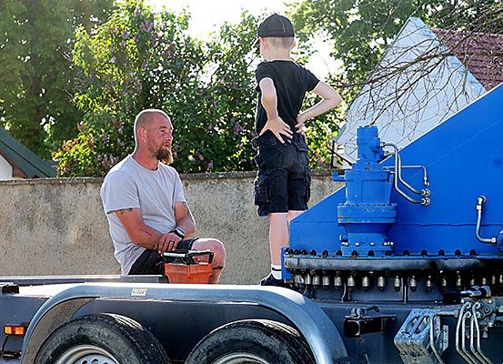 Ein Mann sitzt auf einem Anhänger eines Lastwagens mit einer Werkzeugkiste, während ein Junge an der Seite mit den Händen auf dem Rücken steht, in einem Garten mit einem Haus und Grünpflanzen im Hintergrund.