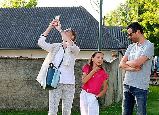 Eine Frau macht ein Foto mit ihrem Handy, ein Mann steht mit verschränkten Armen und ein Mädchen isst etwas. Hinter ihnen steht eine Steinmauer und ein Haus.
