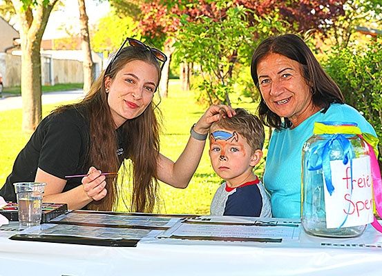 Eine Frau mit Sonnenbrille auf dem Kopf malt das Gesicht eines Kindes mit Farbe. Daneben steht eine weitere lächelnde Frau und ein Glas mit der Aufschrift 'frei Spe'.