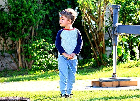 Ein junger Junge mit Gesichtsbemalung steht in einem Garten und schaut zur Seite. Hinter ihm befindet sich eine Holzkonstruktion.