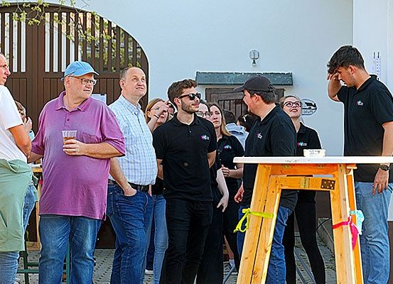 Eine Gruppe von Menschen, einige mit Brille, stehen um einen Tisch herum, wahrscheinlich bei einer Veranstaltung. Sie sind lässig gekleidet und scheinen in ein Gespräch vertieft zu sein.