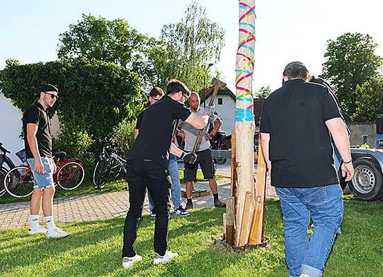 Männer benutzen Hämmer, um einen mit Bändern dekorierten Holzpfahl zu treffen, während ein anderer Mann zusieht. Fahrräder sind an der Seite geparkt.