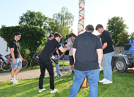 Eine Gruppe von Männern versammelt sich in einem Park. Ein Mann schlägt mit einem Hammer auf einen Pfahl, während andere zusehen. Fahrräder und ein Baufahrzeug sind im Hintergrund.