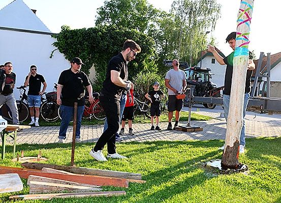 Eine Gruppe von Menschen ist draußen, einer von ihnen schlägt mit einem Hammer auf einen Holzpfahl. Ein Kind steht vor dem Pfahl.