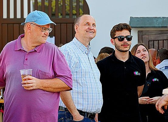Eine Gruppe von Menschen steht zusammen draußen, möglicherweise bei einer Veranstaltung. Ein Mann hält ein Getränk und trägt ein lila Hemd.