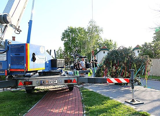 Ein Kran mit Anhänger steht auf einem Ziegelweg. In der Nähe versammeln sich Menschen um einen Baum, möglicherweise zum Beschneiden. Die Umgebung umfasst Häuser, Grünflächen und eine Straße.
