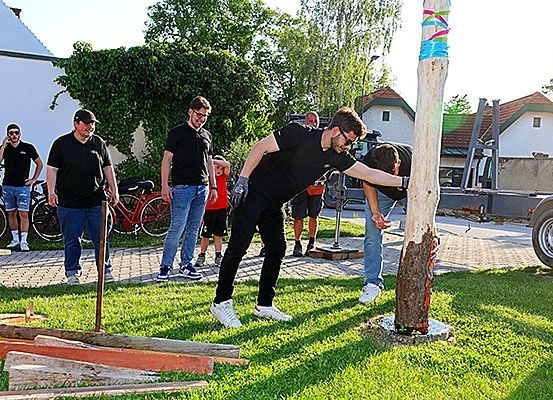 Eine Gruppe von Männern in schwarzen T-Shirts ist draußen, ein Mann nagelt einen Holzpfosten, während andere zusehen und reden. Fahrräder und ein Lastwagen sind in der Nähe.