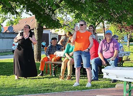 Eine Gruppe von Menschen sitzt auf Bänken unter einem Baum in einem Park. Eine Frau in Schwarz geht und telefoniert. Ein Mann in Orange sitzt mit einer Frau in Grün.