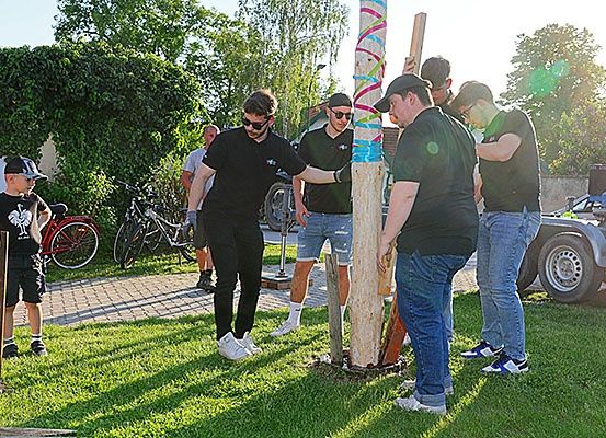 Eine Gruppe von Menschen arbeitet an einem Pfosten in einem Grasbereich. Ein junger Junge steht in der Nähe. Fahrräder und ein Auto sind dahinter geparkt.