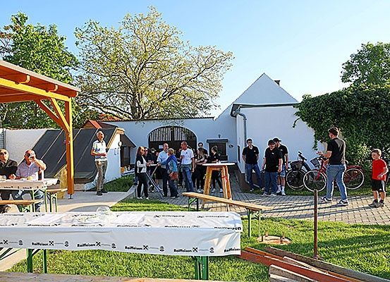 Menschen versammeln sich um Tische im Freien, einige sitzen und andere stehen, in einem Gartenbereich mit geparkten Fahrrädern in der Nähe. Ein Mann steht auf einem Ziegelweg.