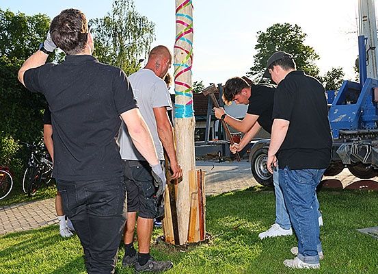 Eine Gruppe von Männern in schwarzen Shirts und Handschuhen arbeitet mit einem Hammer an einem Pfahl, einer trägt einen Hut. Dahinter steht ein blaues Maschinenfahrzeug.