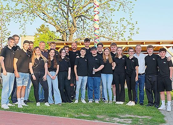 Eine Gruppe junger Menschen in schwarzen T-Shirts und Jeans posiert für ein Foto auf einem Grasfeld mit einem hölzernen Pavillon im Hintergrund.