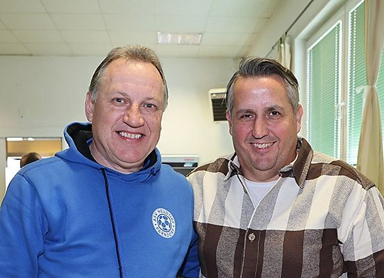 Zwei Männer lächeln in die Kamera, einer in einer blauen Kapuzenjacke und der andere in einem gestreiften Hemd, stehen nebeneinander in einem Raum.