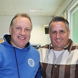 Zwei Männer lächeln in die Kamera, einer in einer blauen Kapuzenjacke und der andere in einem gestreiften Hemd, stehen nebeneinander in einem Raum.