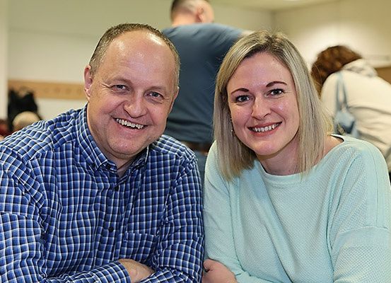 Ein Mann und eine Frau lächeln und posieren für ein Foto in einem Raum mit anderen Personen im Hintergrund.