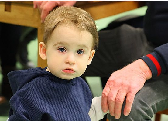Ein junger Junge in einer blauen Kapuzenjacke schaut zur Seite, während er neben einem Erwachsenen steht, der seine Hand hält. Dahinter deutet ein Holztisch und ein unscharfer Hintergrund auf einen Raum oder Saal hin.