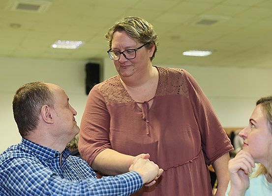 Eine Frau mit Brille steht zwischen zwei Personen, von denen eine ihre Hand hält, während die andere sie ansieht. Die Frau trägt ein kastanienbraunes Oberteil.