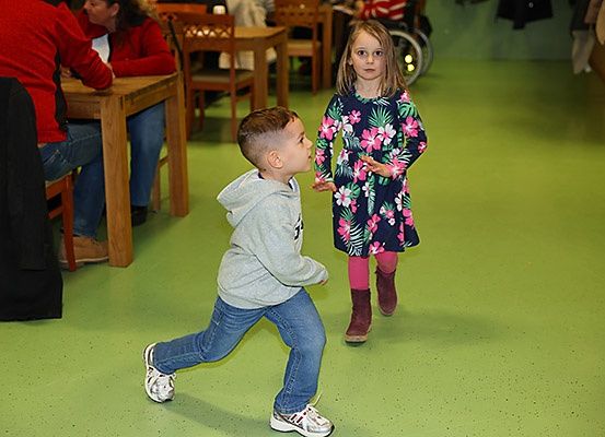 Ein Junge und ein Mädchen gehen in einem Raum mit grünem Boden. Der Junge trägt Turnschuhe und das Mädchen Stiefel. Sie schauen sich an. Hinter ihnen sitzen Leute an einem Tisch.