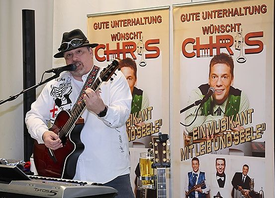 Ein Mann spielt Gitarre und Keyboard vor einem Banner, das 'Chris' mit einem lächelnden Musiker darauf bewirbt.