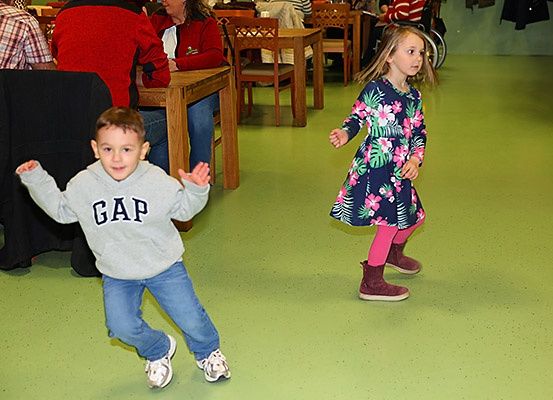 Zwei Kinder tanzen in einem Raum mit grünem Boden, während andere am Tisch sitzen.