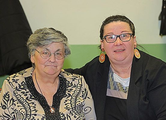 Zwei Frauen sitzen nebeneinander, eine trägt eine Brille und eine Halskette, die andere trägt einen Blazer und Ohrringe. Sie lächeln für ein Foto.