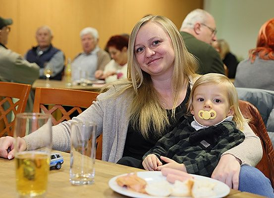 Eine Frau hält ein Kind mit einem Schnuller in einem Essbereich, in dem sich mehrere Personen befinden, darunter ein Mann und eine Frau mit einem Bierglas und einem Essensteller vor ihnen.