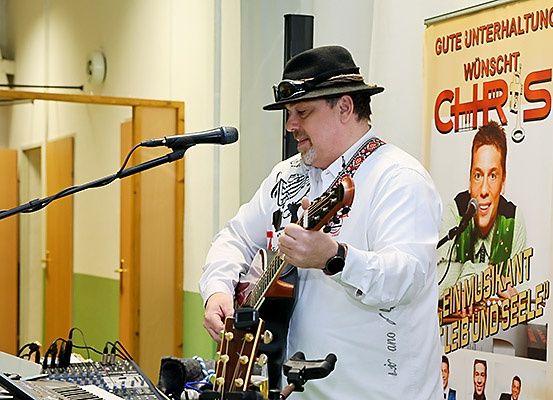 Ein Mann in einem Hut spielt Gitarre vor einem Mikrofon. Hinter ihm steht ein Banner mit dem Wort 'Chris'.