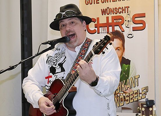 Ein Mann in einem weißen Langarmshirt und einem schwarzen Hut spielt eine Gitarre in ein Mikrofon, mit einem Poster im Hintergrund, das einen Mann und den Text 'Gutes Unwetter Wunscht Chris' zeigt.