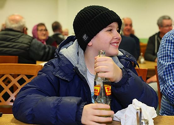 Ein junger Junge, der eine schwarze Mütze und eine blaue gepolsterte Jacke trägt, lächelt, während er eine Glasflasche mit gelbem Etikett hält. Er sitzt an einem Tisch mit anderen in einem Essbereich.