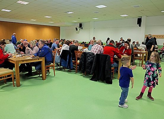 Ein Speisesaal mit Holztischen und Stühlen. Die Leute sitzen und einige stehen, einschließlich eines kleinen Jungen und eines Mädchens. Der Raum hat grünen Boden und weiße Wände.