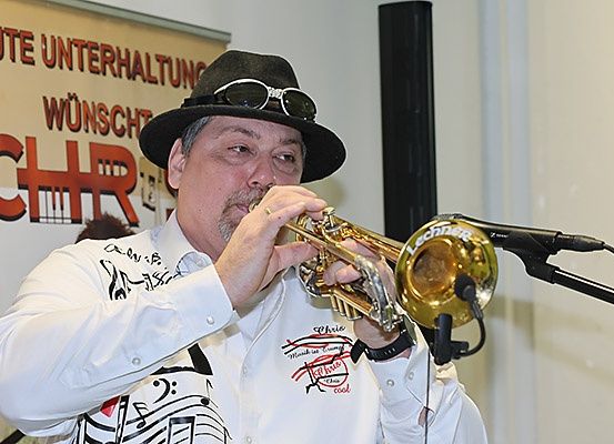 Ein Mann in einem weißen Hemd und schwarzem Hut spielt Trompete vor einem Mikrofon, mit einem Banner, das 'CHR' und 'Gute Unterhaltung Wünscht' im Hintergrund zeigt.