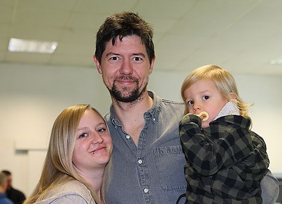 Ein Mann, eine Frau und ein junges Kind lächeln und posieren für ein Foto. Der Mann hat einen Bart und trägt ein graues Hemd. Die Frau hat blonde Haare und trägt eine graue Jacke. Das Kind trägt eine schwarz-weiße Jacke und hält einen Schnuller.