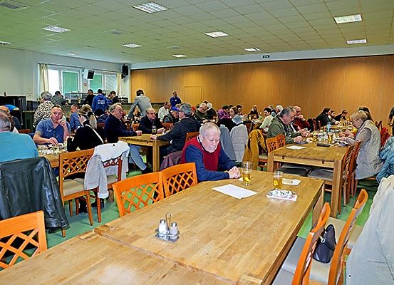 Innenspeisesaal mit Holztischen und Stühlen, eine Gruppe von Menschen sitzt und unterhält sich, Getränke und Essen auf den Tischen, eine Frau steht in der Nähe der Wand.