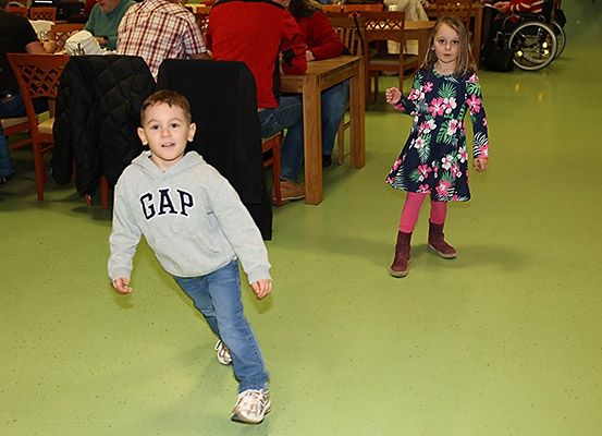 Zwei Kinder, ein Junge und ein Mädchen, laufen gemeinsam in einem Raum mit grünem Boden. Hinter ihnen sitzen Menschen an Tischen.