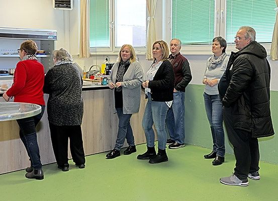 Eine Gruppe von Menschen steht in einem Küchenbereich, einige in der Nähe einer Theke und andere in einer Reihe, im Gespräch. Fenster mit Jalousien und ein Kühlschrank sind im Hintergrund zu sehen.