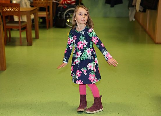 Ein kleines Mädchen in einem Blumenkleid und rosa Strumpfhosen steht in einem grünen Raum, im Hintergrund ist ein Rollstuhl zu sehen.