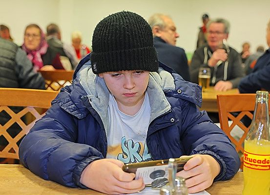Ein junger Junge mit schwarzer Mütze sitzt am Tisch, vertieft in sein Handy, umgeben von Erwachsenen in einem Essbereich, mit einem Glas Bier und einer Flasche Trucade in der Nähe.