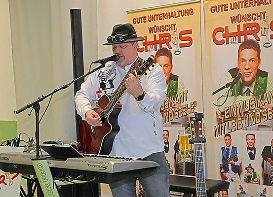 Ein Mann in Hut und Sonnenbrille spielt Gitarre und Keyboard und singt in ein Mikrofon auf der Bühne. Ein Plakat mit 'CHR' und einem Mann im Anzug steht dahinter.