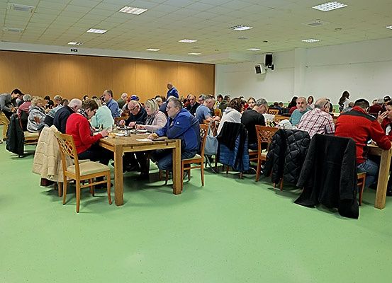Eine große Gruppe von Menschen sitzt in einem Raum an Tischen und trägt Winterkleidung. Der Raum hat einen grünen Boden und eine Holzwand, mit Stühlen und Jacken um die Tische herum.