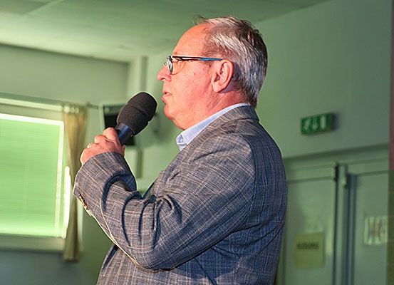 Ein älterer Mann in Anzug und Brille spricht in ein Mikrofon in einem Raum mit grüner Beleuchtung und einem Fenster.