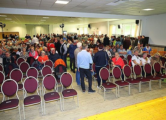 Ein Saal voller Menschen, die auf Stühlen sitzen, einige stehen, mit Deckenleuchten und Fenstern mit Vorhängen.