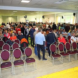 Ein Saal voller Menschen, die auf Stühlen sitzen, einige stehen, mit Deckenleuchten und Fenstern mit Vorhängen.