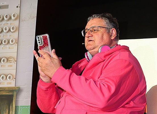 Ein Mann mit Brille und einer rosa Kapuzenjacke hält ein Smartphone und scheint ein Foto zu machen.