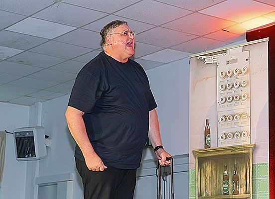 Ein Mann steht auf der Bühne, singt in ein Mikrofon, mit einem Regal mit Flaschen und einem Deckenventilator im Hintergrund.