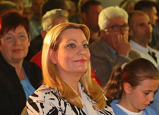 Eine Frau mit blonden Haaren sitzt inmitten einer Menschenmenge. Hinter ihr schaut eine Frau mit roten Haaren und Brille zur Seite. Eine junge Mädchen mit einem Pferdeschwanz sitzt zu ihrer Rechten.